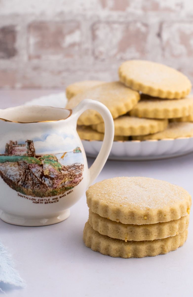 A decorative jug with a scenic print of Tantallon Castle sits beside a stack of three sugar-dusted lemon shortbread cookies - make to the traditional recipe for Tantallon Cakes. More biscuits, are arranged on a plate in the background, all on a light-coloured surface.