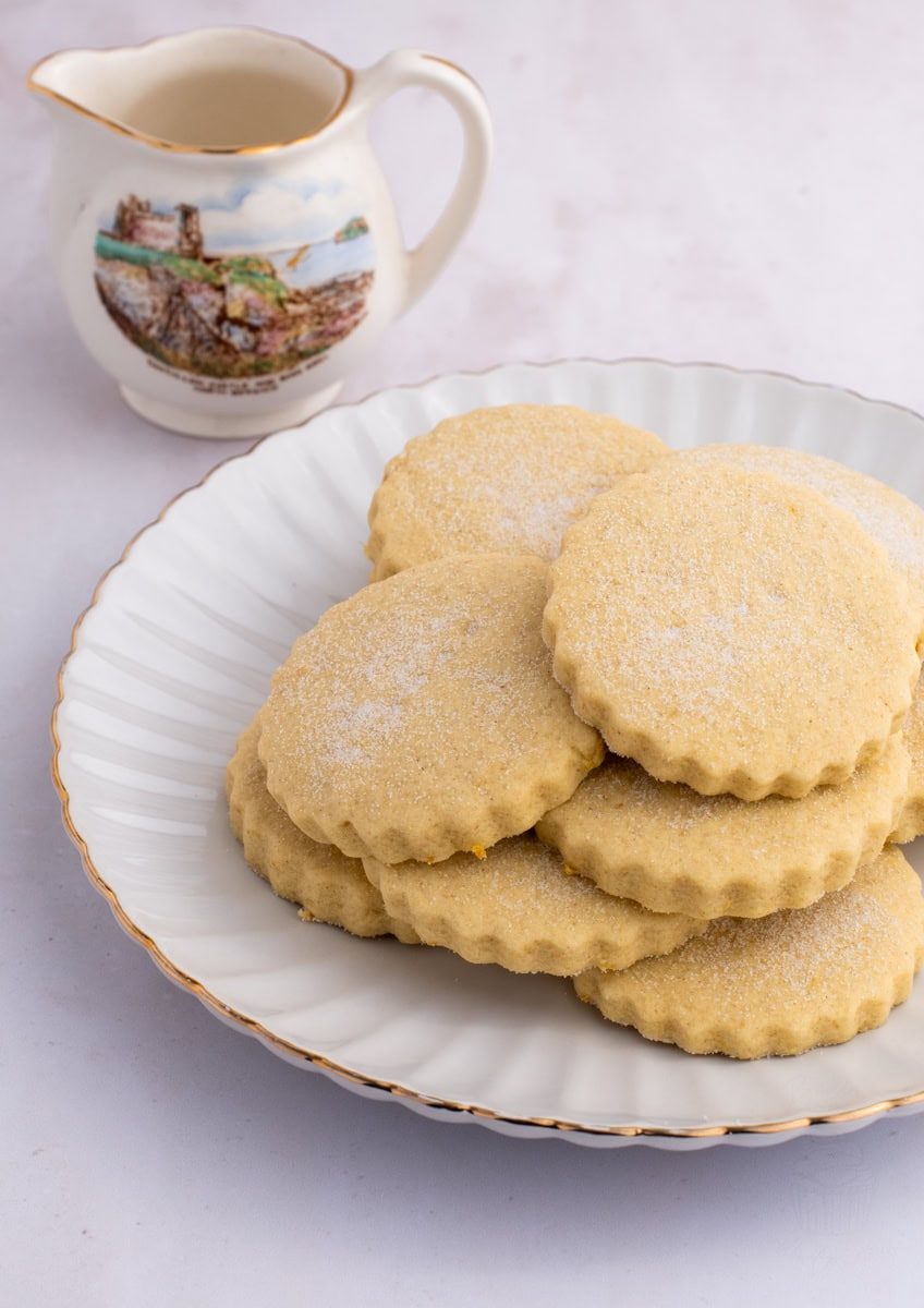 A white plate holds several sugar-dusted shortbread cookies, reminiscent of traditional Scottish Tantallon cakes. In the background, there's a ceramic jug with a scenic painting of Tantallon Castle on it. The light-coloured surface adds a soft contrast to the delightful cookies.