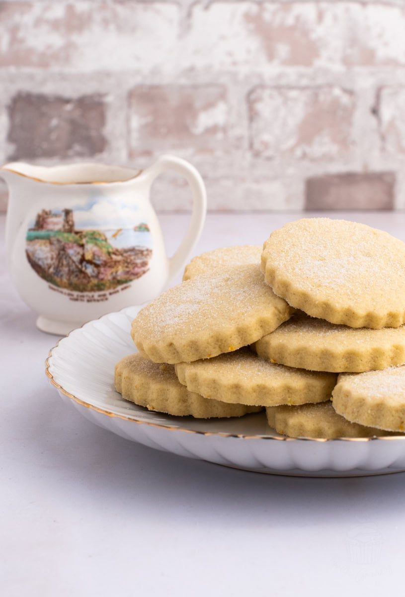 A plate of sugar-dusted shortbread cookies reminiscent of traditional Tantallon Cakes rests elegantly on a white, scalloped-edge plate. Behind it, a decorative cream jug featuring a watercolor landscape of Tantallon Castle sits gracefully on a white surface, with a blurred brick wall in the background.