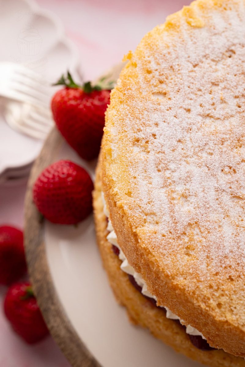 Victoria Sponge Top-down view of a Victoria Sponge cake dusted with caster sugar, surrounded by strawberries on a rustic cake stand.